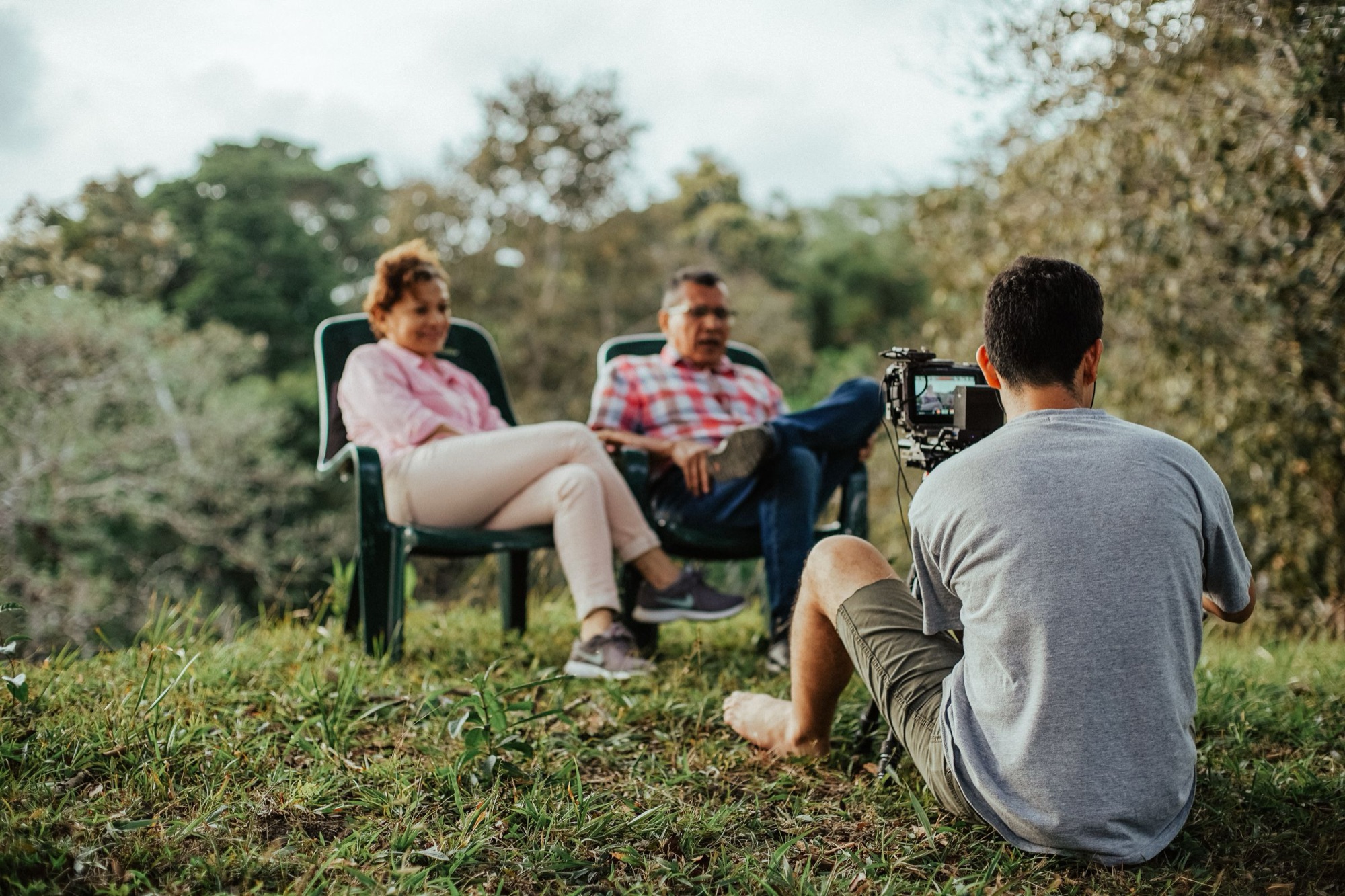 A camera operator filming a couple sitting in garden chairs