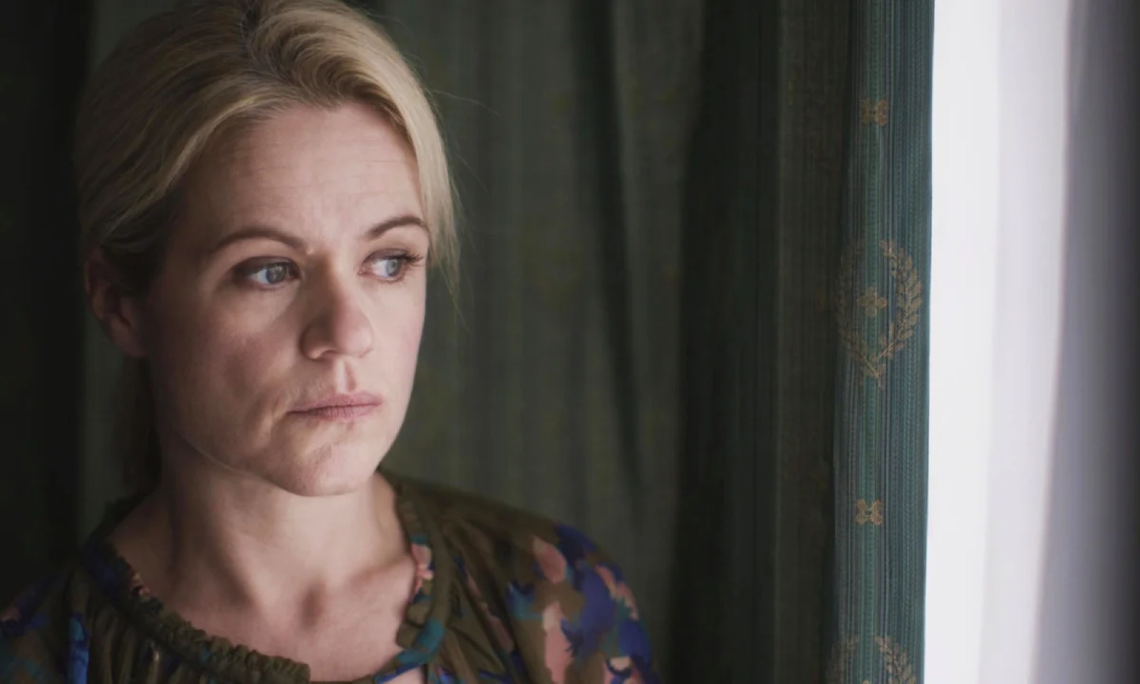 A woman looks out of a window beside a green curtain