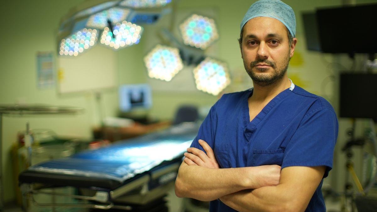 A surgeon in blue scrubs stands with arms folded in an operating theatre